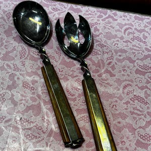 May include: A pair of salad serving utensils featuring polished silver-tone metal heads and twisted necks. The handles are constructed from dark brown wood with a hexagonal shape, providing a sturdy grip for these functional kitchen tools.