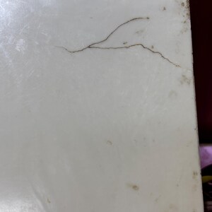 May include: Close-up of a white, rectangular surface with a prominent brown crack running across it. The surface has minor discoloration and imperfections, suggesting age or wear. The edges show slight staining.