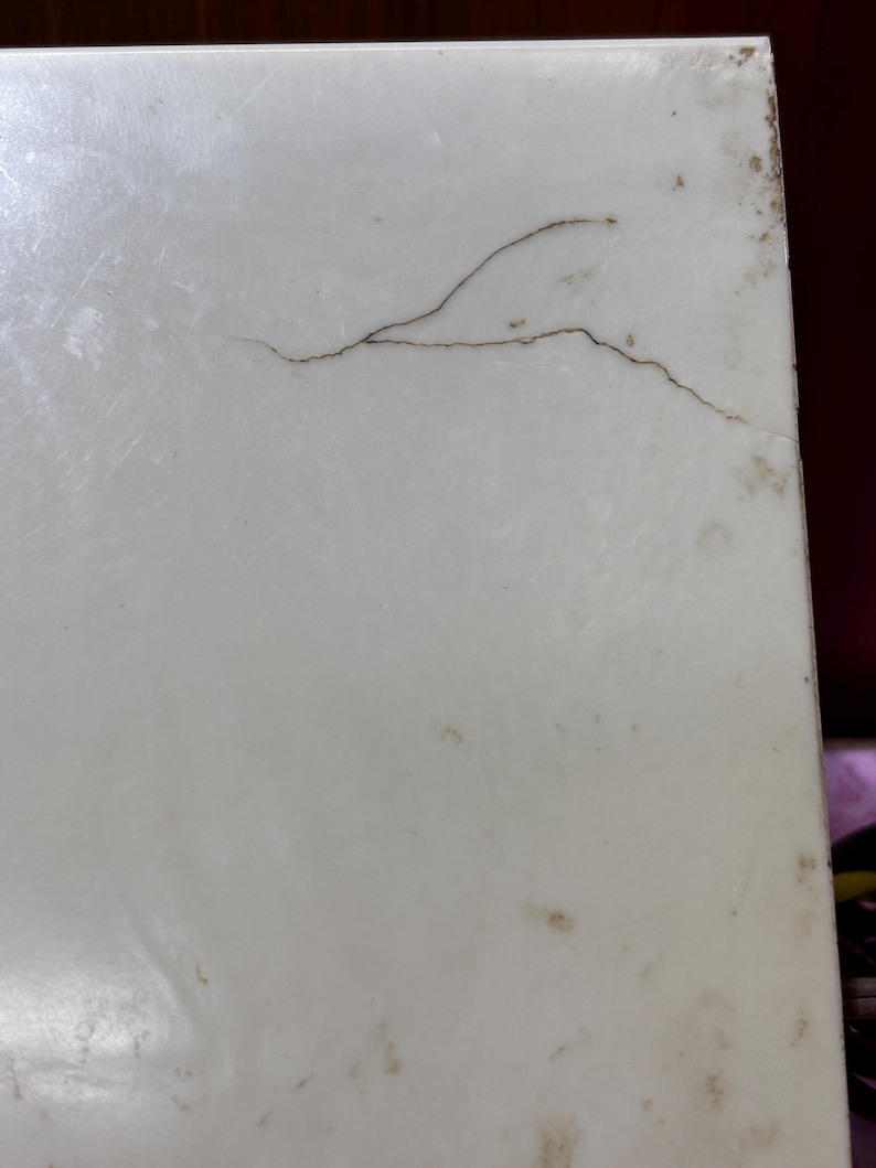 May include: Close-up of a white, rectangular surface with a prominent brown crack running across it. The surface has minor discoloration and imperfections, suggesting age or wear. The edges show slight staining.