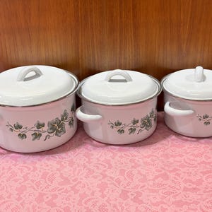 May include: A set of three white enamel pots with lids, featuring a silver rim and handles. Each pot is decorated with a floral vine design in shades of gray and green. The pots are arranged on a pink lace tablecloth.