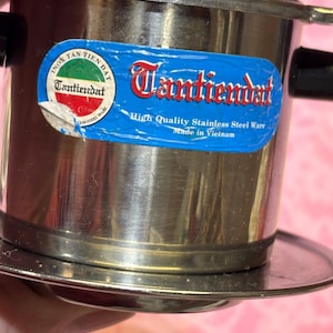 May include: Stainless steel Vietnamese coffee filter with the text "Tantiendat" on a blue sticker. The filter has a round base and a cylindrical body with black handles. The sticker also says "High Quality Stainless Steel Ware Made in Vietnam."