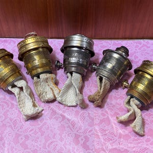 May include: Five vintage oil lamp burners. The burners are made of brass and silver-colored metal, with a perforated design. Each burner has a wick extending from the bottom. The background is a pink floral pattern.