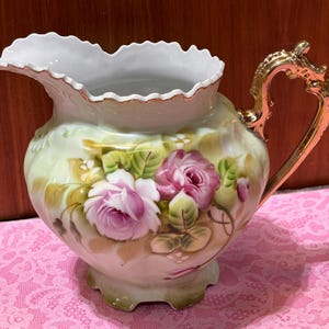 May include: A porcelain creamer pitcher featuring a scalloped rim and a gold-toned handle. The body is decorated with hand-painted pink roses and green foliage against a pale cream background, finished with a gold-trimmed base.