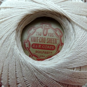 May include: A close-up of a ball of off-white "KNIT-CRO-SHEEN" thread. The label reads "250 YDS." and "J. & P. COATS BOILFAST U.S.A." The thread is tightly wound, showing the texture of the yarn. The background is a lace doily.