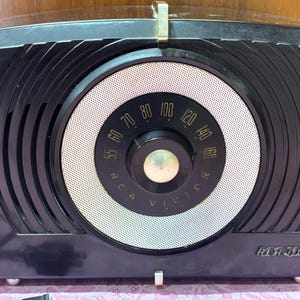 May include: A black RCA Victor tabletop radio featuring a large circular silver speaker grille, a central black tuning dial with gold numbers ranging from 55 to 160, and a gold-colored RCA Victor logo printed on the lower right corner.