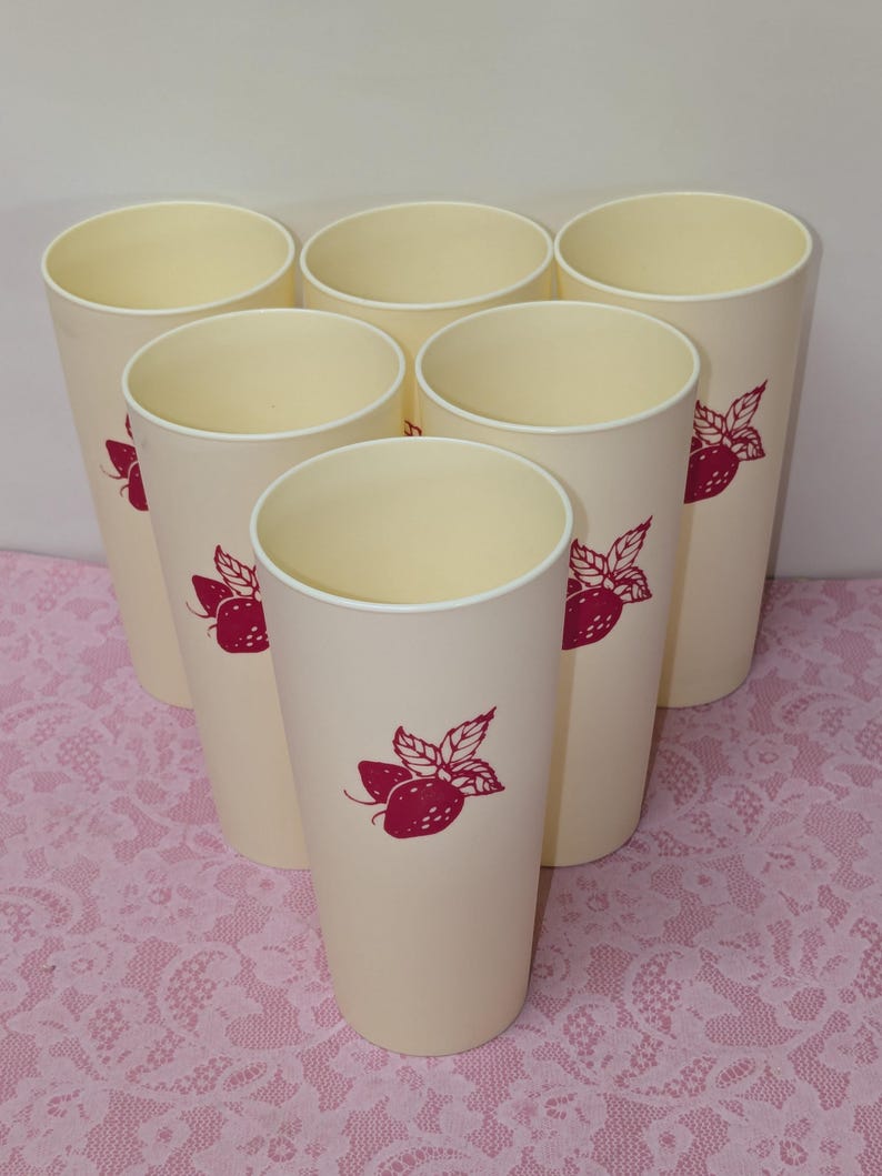 May include: Set of seven cream-colored tall plastic tumblers. Each tumbler features a red strawberry design. The tumblers are arranged in a semi-circular pattern, showcasing their uniform shape and design. Ideal for serving cold beverages.