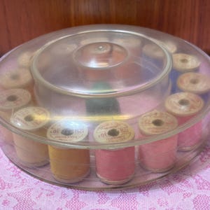 May include: A clear plastic sewing thread organizer filled with spools of thread in shades of pink and yellow. The spools are arranged in a circular pattern inside the container. The thread spools have a label that reads "30".