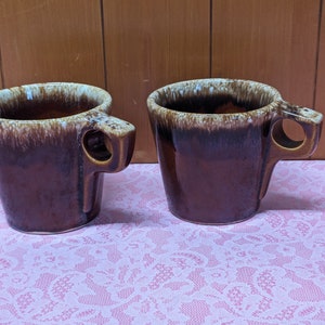 May include: Two brown ceramic mugs with a unique handle design. The mugs have a dark brown exterior with a lighter brown and white rim. The handles feature a circular opening for easy gripping. The mugs are placed on a pink lace tablecloth.