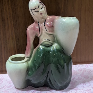 May include: Vintage ceramic figurine of a woman in a long green skirt and pink top, holding two light green vases. The figurine has a detailed face and is likely a decorative piece.