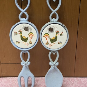 May include: A pair of blue cast iron pot holders with a rooster and sun design on the front. The pot holders are shaped like a fork and a spoon.