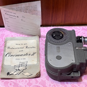 May include: Vintage Cinemaster II dual 8mm movie camera with a registration card and instruction booklet. The camera is gray and black with a lens and dials. The booklet has the title "How to get Professional Results with Cinemaster II".
