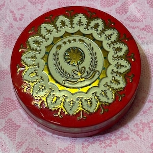 May include: A round, red tin with a decorative lid. The lid features a central design with a gold starburst, surrounded by a white and gold floral pattern. The tin's edges are also adorned with gold accents.
