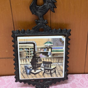 May include: Black cast iron pot holder with a rooster design. The holder has a square tile with a vintage illustration of a general store interior with a wood stove, a chair, and a bucket.
