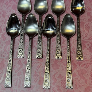 May include: A set of eight silver spoons. Each spoon has a rounded bowl and a long handle with a floral design. The spoons are arranged on a pink patterned surface.