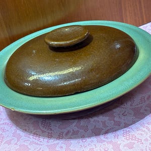 May include: An oval-shaped, brown ceramic casserole dish with a matching lid and a light green rim. The lid has a small, round handle. The dish sits on a pink and white patterned surface.