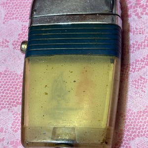 May include: Vintage lighter with a silver metal top and a clear, yellowed plastic body. The lighter has a dark blue band around the middle and a small, round button on the side. The lighter shows signs of wear and age.