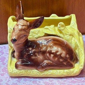 May include: A ceramic planter featuring a brown fawn resting in front of a bright yellow textured log. The fawn has dark brown fur with lighter spots on its back and a black nose. The planter has an open top and a glossy finish throughout.