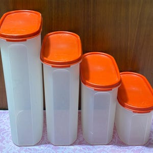 May include: A set of four plastic food storage containers. The containers are white with orange lids. The containers are different sizes, ranging from tall and slender to short and wide. The lids are oval-shaped and fit snugly on top of the containers.