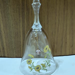 May include: Clear glass bell with a floral design featuring yellow sunflowers and green leaves. A small pink crystal hangs from a gold chain inside the bell. The bell has a long, slender handle and a flared base.