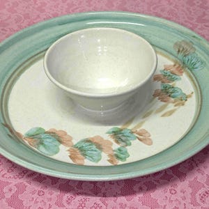 May include: A ceramic serving set featuring a round bowl nested on a large oval platter. The set is primarily off-white with a teal rim and floral accents in teal and peach. The set is designed for serving food.