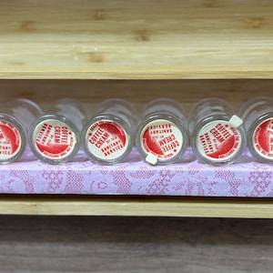 May include: A row of vintage glass coffee cream bottles with red and white labels. The labels read "Coffee Cream Sanitary Service". The bottles are arranged on a pink lace-patterned shelf inside a wooden box.