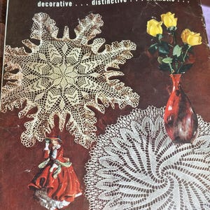 May include: Vintage book cover featuring the word "DOILIES" in yellow lettering. The cover showcases examples of doilies, a red vase with yellow roses, and a figurine. The doilies are white and intricately designed.