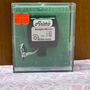 May include: A vintage digital pedometer in a clear plastic case. The pedometer is black with a white display and the brand name "Aristo". The case has a green backing and a price tag from "Spike's Sporting Goods". The pedometer measures in feet.