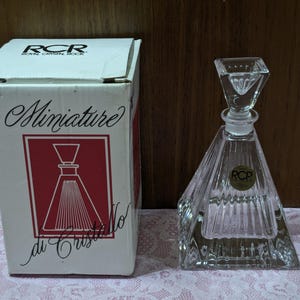 May include: A clear, faceted crystal perfume bottle with a geometric design, next to its original box. The box is white with red accents and the words "Miniature di Cristallo". The bottle has a square stopper.