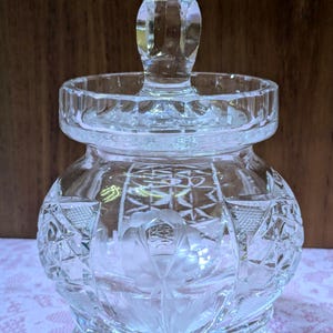 May include: A clear, cut-glass jar with a lid and three small feet. The jar features intricate geometric and floral patterns, with a faceted knob on the lid. The jar is designed to hold small items.