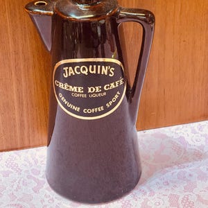 May include: A dark brown ceramic Jacquin's Crème de Café coffee liqueur bottle with a black cap and handle. The bottle has a gold label with black text that reads "Jacquin's Crème de Café Coffee Liqueur Genuine Coffee Sport."