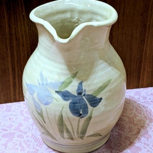 May include: A handmade ceramic pitcher with a cream-colored glaze. The pitcher is adorned with hand-painted blue iris flowers and green leaves. The pitcher has a rounded body and a spout for pouring.