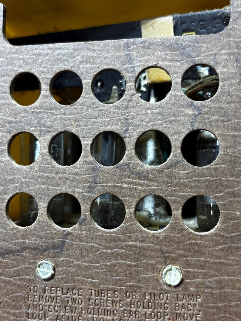 May include: Close-up of a vintage radio component with a brown, textured surface. The component features multiple circular openings revealing internal parts. Text at the bottom instructs on tube replacement. Two screws are visible.