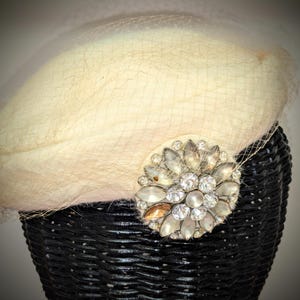 May include: Cream-colored vintage hat with a delicate veil and a sparkling rhinestone brooch. The hat is set on a black wicker form, showcasing its elegant design. A classic accessory for special occasions.
