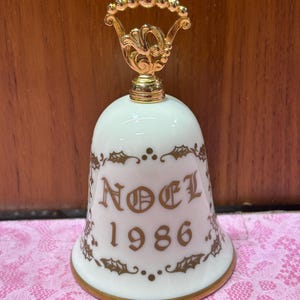 May include: A white porcelain bell featuring a gold-colored handle with a decorative filigree design. The body of the bell is adorned with gold-colored holly leaf patterns and the text NOEL 1986 printed in a gold-colored serif font on the front.