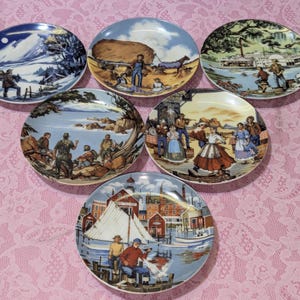 May include: A set of six decorative plates with colorful scenes. Each plate features a different vintage illustration, including landscapes, people, and historical events. The plates are arranged on a pink lace tablecloth.