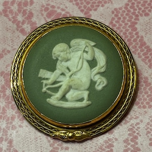 May include: A round, antique brooch with a gold-tone frame. The center features a green background with a white relief of a cherub with a bow and arrow. The brooch has a detailed, decorative border.