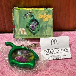 May include: A green "a bug's life" keychain watch from McDonald's. The watch has a green frame with a picture of the characters from the movie. The watch is on a pink surface with a "Clip-Tock Watch Collection" card.