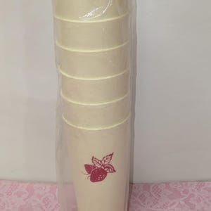 May include: A set of six cream-colored plastic tumblers, wrapped in clear plastic. Each tumbler has a horizontal band design and a red strawberry graphic. The tumblers are cylindrical and appear to be new.