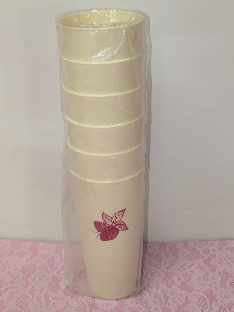 May include: A set of six cream-colored plastic tumblers, wrapped in clear plastic. Each tumbler has a horizontal band design and a red strawberry graphic. The tumblers are cylindrical and appear to be new.