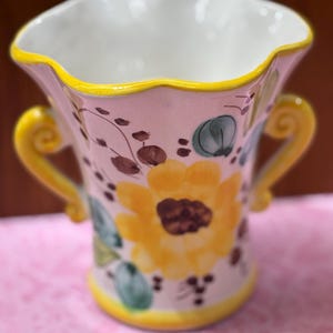May include: A ceramic vase with a ruffled rim, yellow handles, and a yellow base. The vase is decorated with a large yellow sunflower, green leaves, and brown floral accents. The interior is white.