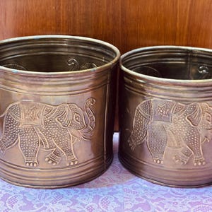 May include: Two antique brass-colored cylindrical planters. Each planter features an embossed elephant design with intricate details. The planters are of similar size and shape, suitable for indoor or outdoor use.