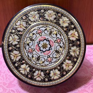 May include: A round metal tin featuring a dark navy blue background with a decorative border of white flowers with yellow centers. The center of the lid displays an intricate gold and white filigree pattern with a central star and pink accents.