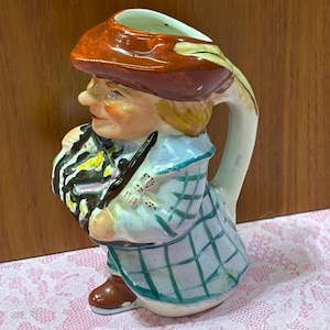 May include: A ceramic pitcher in the shape of a man wearing a brown hat and plaid coat. The pitcher has a white handle and a detailed face. The man is holding a black and white striped object. The pitcher is on a pink lace surface.