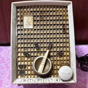 May include: Vintage Emerson radio with a cream-colored frame and a gold-colored speaker grill. The radio features a tuning dial, a volume knob, and the Emerson logo. The radio's design is a classic example of mid-century electronics.