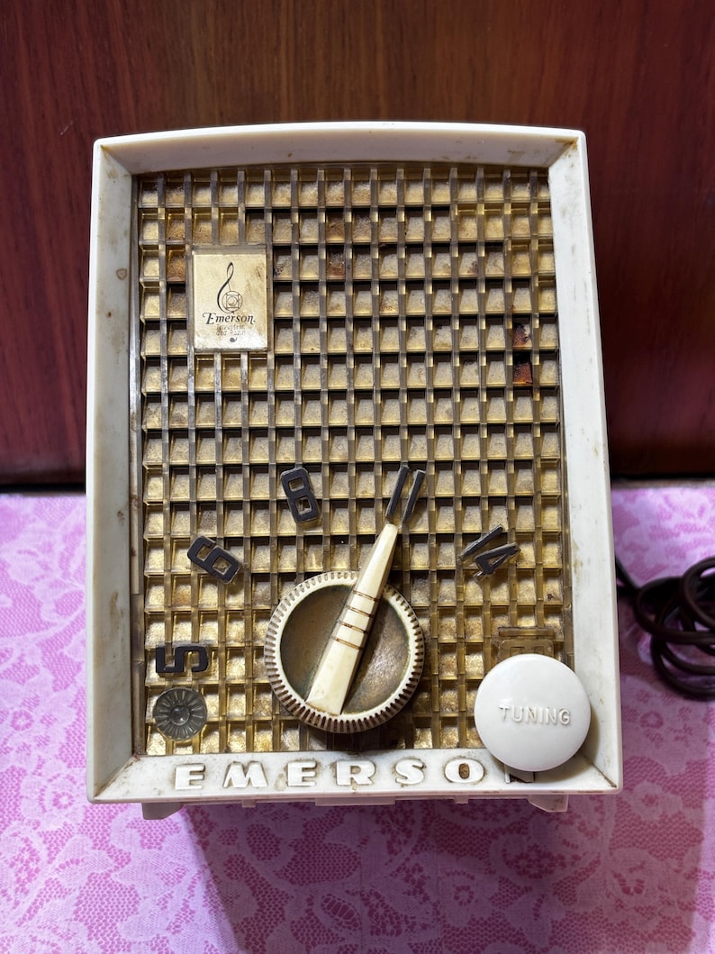 May include: Vintage Emerson radio with a cream-colored frame and a gold-colored speaker grill. The radio features a tuning dial, a volume knob, and the Emerson logo. The radio's design is a classic example of mid-century electronics.