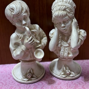 May include: Two off-white ceramic figurines. One is a boy playing a wind instrument, the other is a girl covering her ears. Both figures are on pedestal bases with floral details, and are in a vintage style.