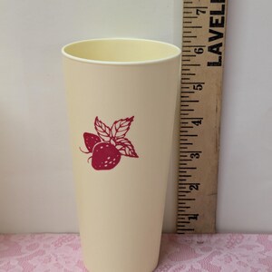 May include: A tall, cream-colored plastic tumbler with a red strawberry design. The tumbler has a slightly flared shape and a smooth surface. A wooden ruler is next to the tumbler.