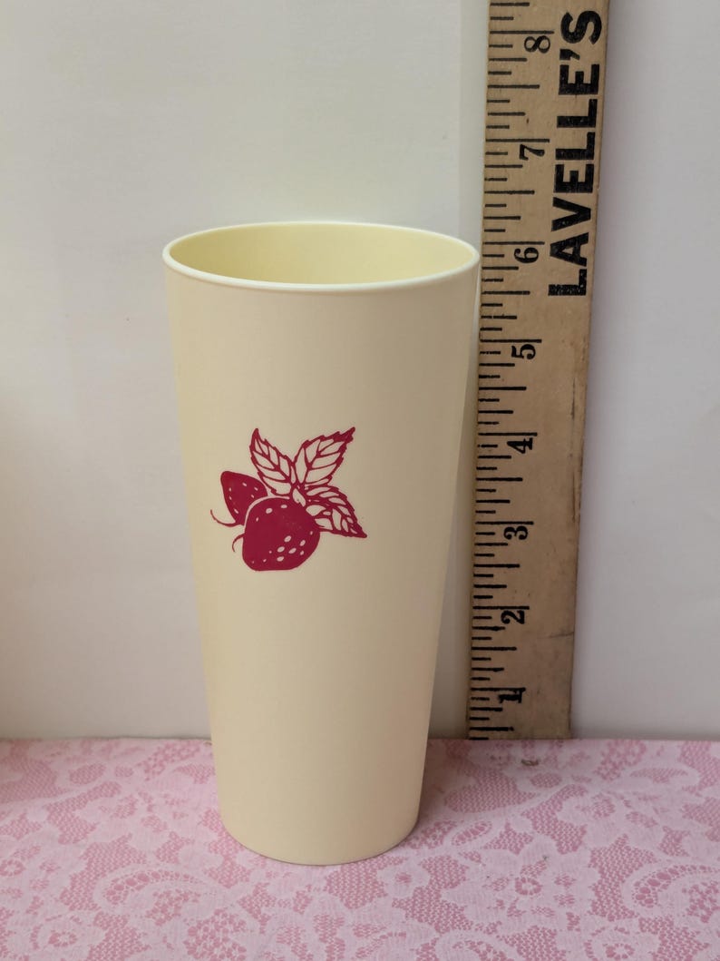May include: A tall, cream-colored plastic tumbler with a red strawberry design. The tumbler has a slightly flared shape and a smooth surface. A wooden ruler is next to the tumbler.