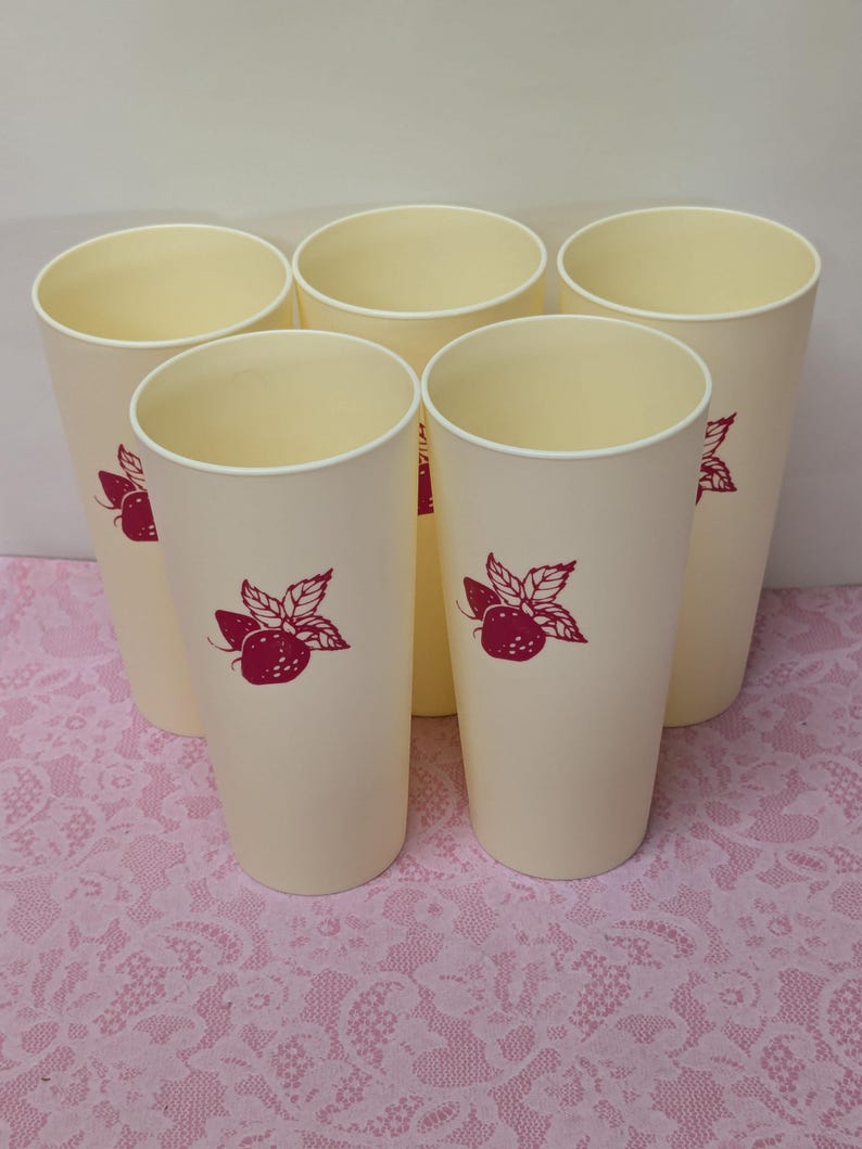 May include: Five tall, cream-colored plastic tumblers with a red strawberry and leaf design. The tumblers have a simple, cylindrical shape and a white rim. The design is repeated on each tumbler.