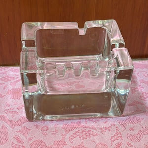 May include: A clear, square glass ashtray with a rectangular shape and four cigarette rests. The ashtray has a stepped design and is made of transparent glass. The ashtray is sitting on a pink lace surface.